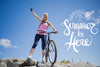 Smiling woman in summer cycling gear standing with her bike on a rocky hill under a blue sky, raising her arm in celebration. Text overlay says 